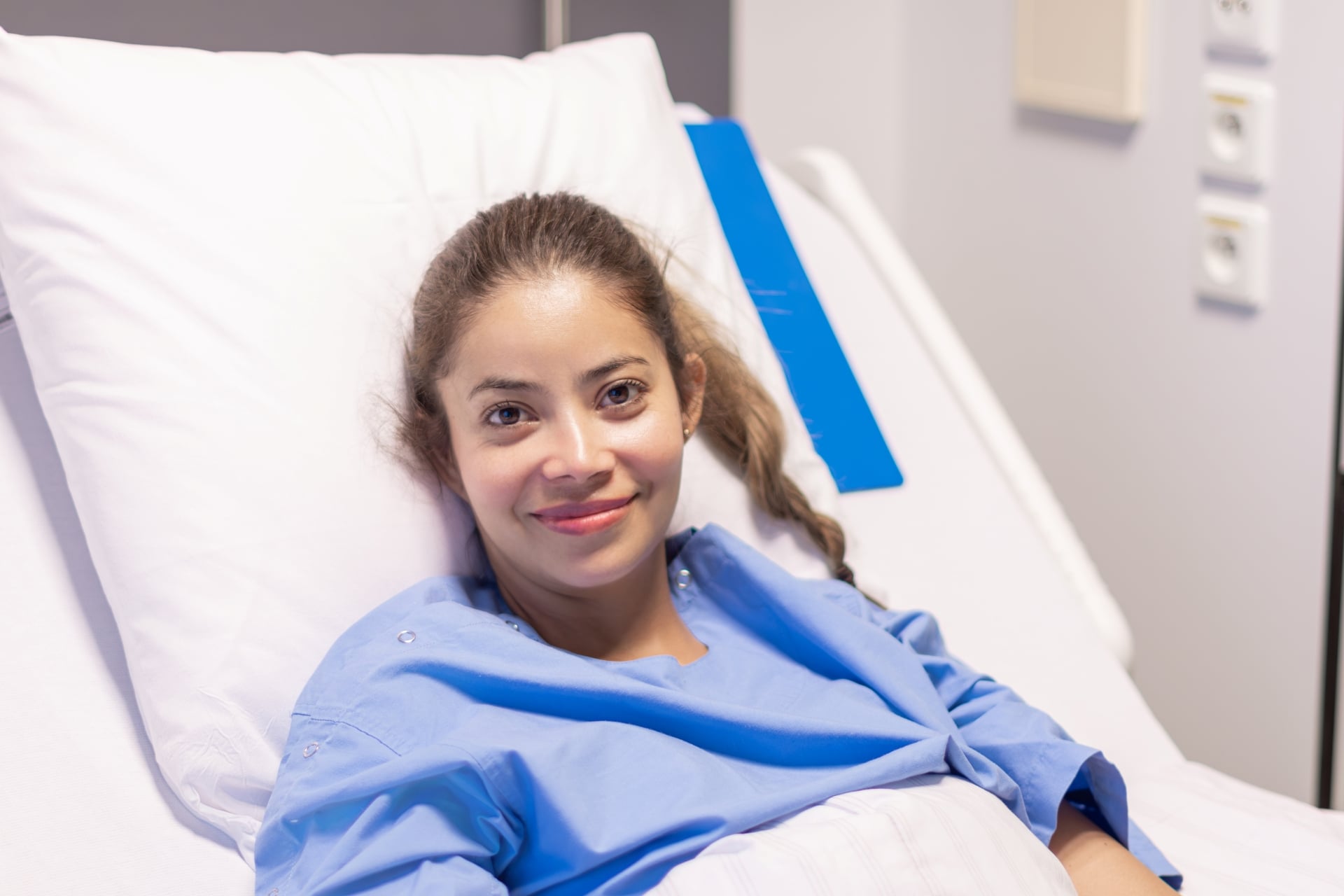 Smiling female patient resting comfortably in a hospital bed, wearing a blue gown after a medical procedure.