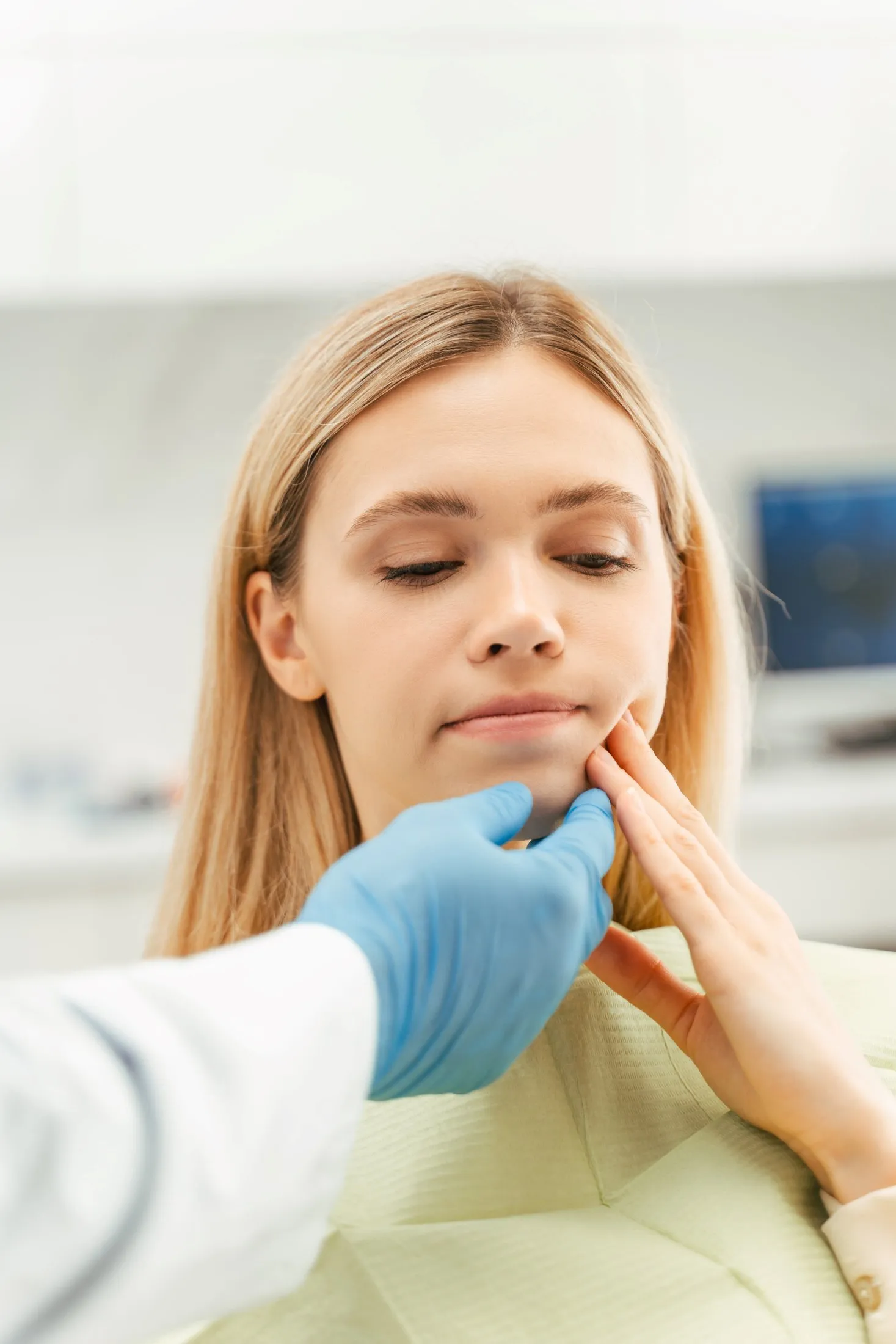 Patient gently holding her cheek while an oral surgeon evaluates jaw alignment and facial swelling in a clinical setting.