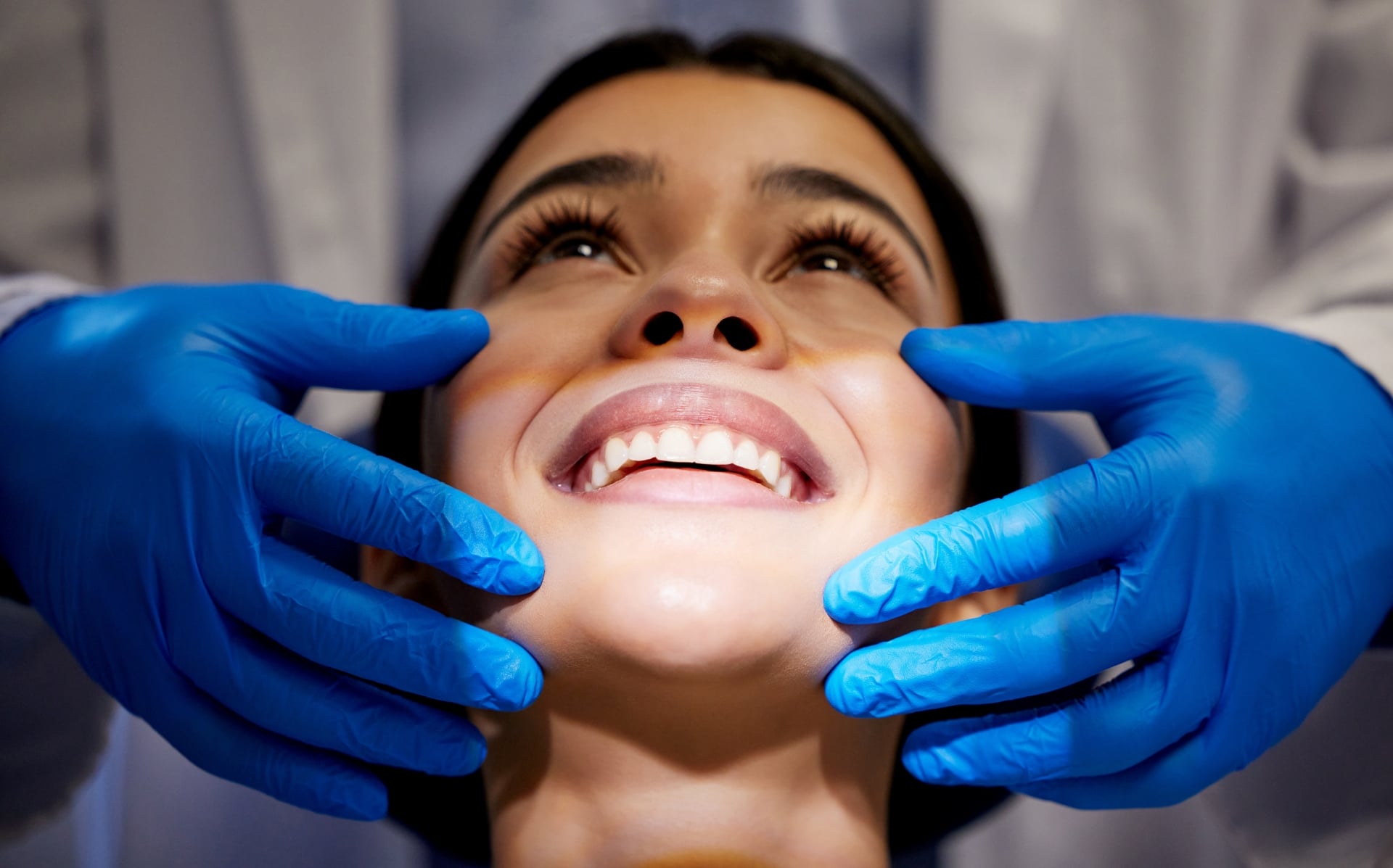 Close-up of smiling woman during dental treatment with blue-gloved hands gently examining her teeth. 