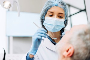 Premier Oral and Maxillofacial Surgery group dentist wearing a mask uses a tool to screen an older male patient.